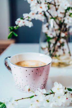 A cup of coffee in a light pink cup and spring blooming branch with flowers on the white table background. Minimalist Vertical card. Soft selective focus, copy space. Tender Spring morning mood