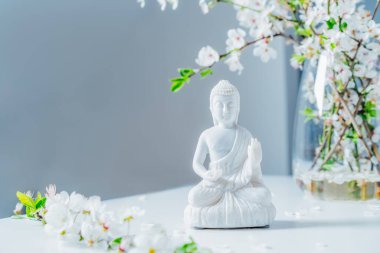 Decorative white Buddha statuette with blooming tree branches in the vase on the white background. Meditation and relaxation ritual. Buddha birthday. Selective focus.