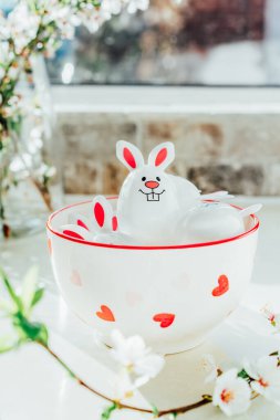 Funny Easter eggs containers with bunny ears filled with candy and chocolate in bowl on white kitchen table with blooming branches. Festive spring Easter composition. Vertical card. Selective focus.