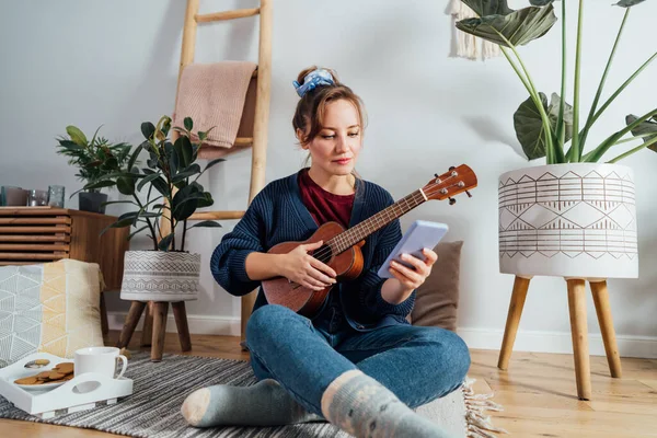 Young Woman using mobile phone while playing ukulele guitar in modern Scandi interior home. Online learning concept. Tutorial lessons via mobile application. New Hobby, resolution to learn new skills