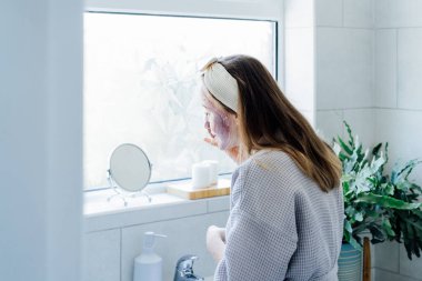Young woman in bathrobe looking in the mirror and removing natural clay mask with loofah pad from face in bathroom. Cosmetic procedures for teens skin care at home. Beauty self-care. Selective focus.
