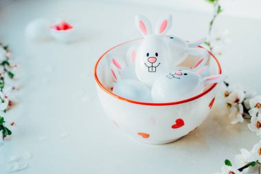 Komik Paskalya yumurtalarını, içi şeker ve çikolata dolu tavşancık kulaklı kapları beyaz mutfak masasında, dallarla dolu bir kasede kapatın. Bayramlık bahar paskalya kompozisyonu. Seçici odak.