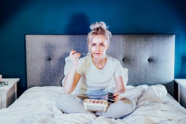 Bored, frustated woman eating ice cream and using the remote control to change the channel while watching television in bed at night. Overeating, Jamming of negative emotions. Selective focus
