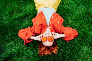 Top view happy smiling redhead Woman with freckles, red orange gerbera Flower glasses lying on green grass. Positive Emotion people. Fashion girl in bright colors jeans clothes. Spring, summer mood