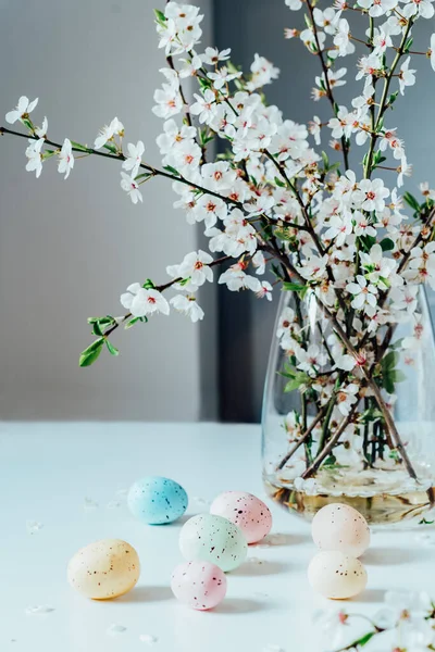 Blooming tree branches in the vase and colored easter eggs on the white kitchen table with simple interior background. Easter celebration at home. Festive spring vertical card. Selective focus