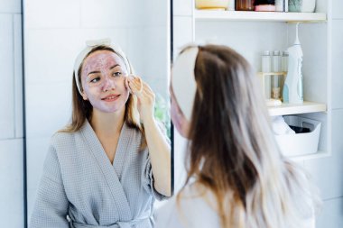 Young woman in bathrobe looking in the mirror and removing natural clay mask with loofah pad from face in bathroom. Cosmetic procedures for teens skin care at home. Beauty self-care. Selective focus.