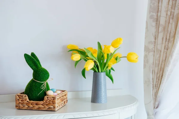 Easter home decor. Easter bunny rabbit statuette in straw basket with colored eggs, fresh spring tulips and daffodils flowers on the chest drawers in light classic interior design. Selective focus