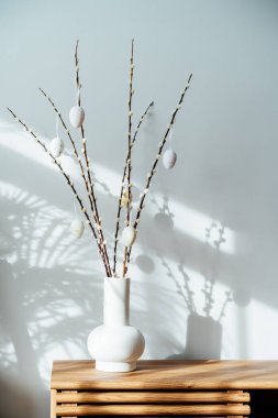 Easter home decor. Modern minimalist style interior with blooming branches of the pussy willow and hanging easter eggs in ceramic vase on a wooden console with sunlight and shadows on white gray wall