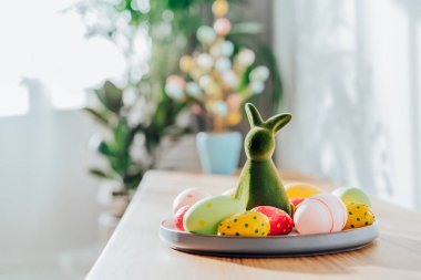 Easter home decor. Green bunny rabbit figurine and colored easter chocolate eggs on the plate and easter tree on wooden console with sunlight and shadows on white wall. Selective focus. Copy space