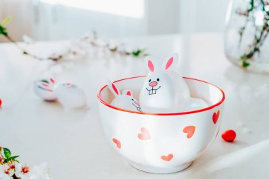 Komik Paskalya yumurtalarını, içi şeker ve çikolata dolu tavşancık kulaklı kapları beyaz mutfak masasında, dallarla dolu bir kasede kapatın. Bayramlık bahar paskalya kompozisyonu. Seçici odak.