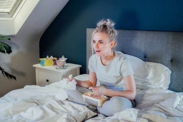 Young excited woman in home clothes passionately watching Tv or movie with bated breath. Girl eating ice cream, holding tissue while sitting on bed at home alone. Leisure, relaxation time.