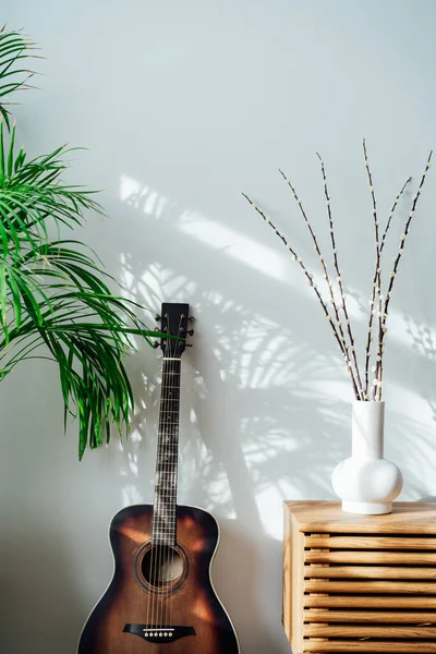 Modern minimalist Scandinavian style interior with branches of the pussy willow on a wooden console, green house plants and acoustic guitar near white wall with shadows on white wall. Vertical card