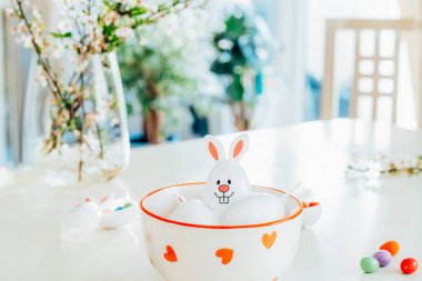 Komik Paskalya yumurtalarını, içi şeker ve çikolata dolu tavşancık kulaklı kapları beyaz mutfak masasında, dallarla dolu bir kasede kapatın. Bayramlık bahar paskalya kompozisyonu. Seçici odak.