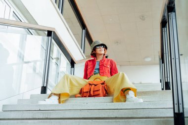 Confident Hipster young woman in bright clothes, sunglasses, backpack bag and bucket hat sitting on the stairs in public place. Urban city street fashion. Fashion blogger. Selective focus. Copy space