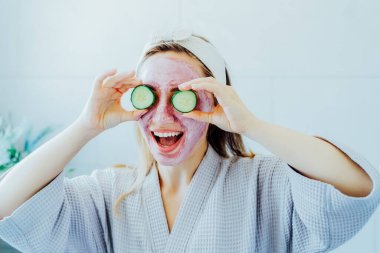 Yüzünde pembe kilden maske olan genç bir kadın banyoda salatalık dilimleri tutuyor ve taze bir maske yapıyor. Evde cilt bakımı için doğal kozmetik prosedürleri. Güzellik özgüveni. Seçici odak