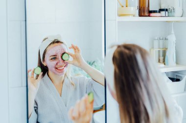 Yüzünde pembe kilden maske olan genç bir kadın banyoda salatalık dilimleri tutuyor ve taze bir maske yapıyor. Evde cilt bakımı için doğal kozmetik prosedürleri. Güzellik özgüveni. Seçici odak
