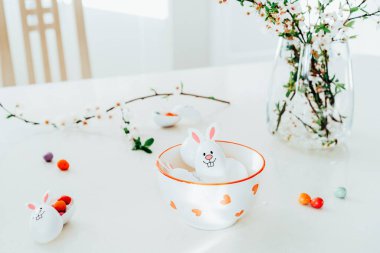 Komik Paskalya yumurtalarını, içi şeker ve çikolata dolu tavşancık kulaklı kapları beyaz mutfak masasında, dallarla dolu bir kasede kapatın. Bayramlık bahar paskalya kompozisyonu. Seçici odak