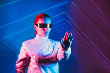 Neon portrait of woman, dressed in futuristic holographic clothes and glasses touch space by fingers. Cyberpunk girl with virtual touching pad. Neon blue and pink light. Virtual reality glasses