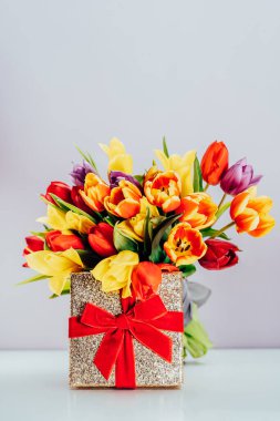 Celebration concept - golden gift box with red velvet ribbon and tulip flowers bouquet. Vertical card mockup. Womens day, 8 march, mothers day, birthday greetings. Selective focus. Copy space.