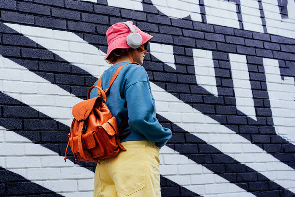 Back view stylish woman in bright clothes and bucket hat wearing wireless headphones and listening to music while walking near city urban painted wall. Fashionable hipster lifestyle. Selective focus.
