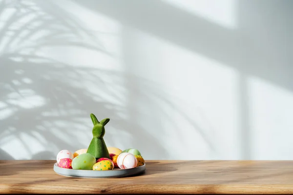 Easter home decor. Composition of Green bunny rabbit figurine and colored easter chocolate eggs on the plate on the wooden console with sunlight and shadows on white wall. Selective focus. Copy space