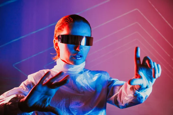 Neon portrait of woman, dressed in futuristic holographic clothes and glasses touch space by fingers. Cyberpunk girl with virtual touching pad. Neon blue and pink light. Virtual reality glasses