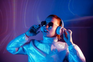 Portrait of dancing woman in sunglasses and headphones in neon light. Music lover. Silent disco. Woman in holographic clothes on the background of music vibes background. Futuristic Party