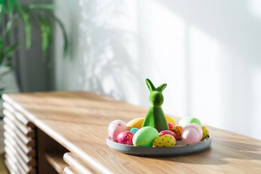 Easter home decor. Green bunny rabbit figurine and colored easter chocolate eggs on the plate and easter tree on wooden console with sunlight and shadows on white wall. Selective focus. Copy space