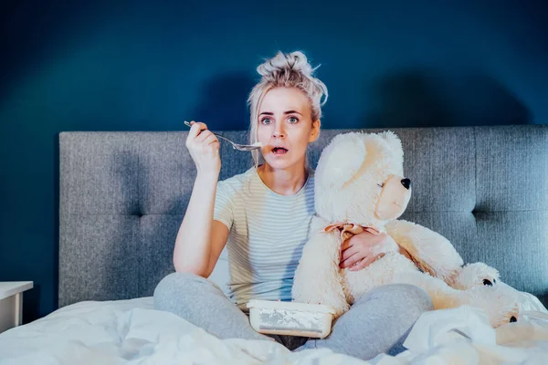 Young woman in home clothes passionately, hugging teddy bear, watching movie with bated breath. Girl eating ice cream while sitting on bed at home in evening alone. Leisure, relaxation time