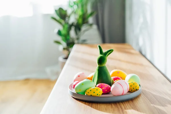 Easter home decor. Green bunny rabbit figurine and colored easter chocolate eggs on the plate and easter tree on wooden console with sunlight and shadows on white wall. Selective focus. Copy space
