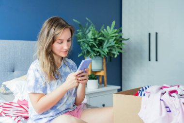 Smiling woman using smartphone to find places where to donate or sale clothes. Decluttering, Sorting clothes and Cleaning Up. Conscious consumer, sustainability. Reuse, second hand, Charity concept.