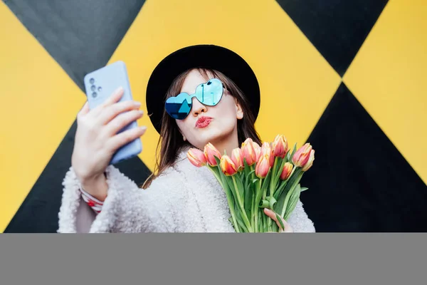 多愁善感的年轻时尚女性戴着心形太阳镜 手持郁金香花束 在几何图形明亮的黄黑色墙壁背景上拍照 城市街道时尚 春天的心情 — 图库照片