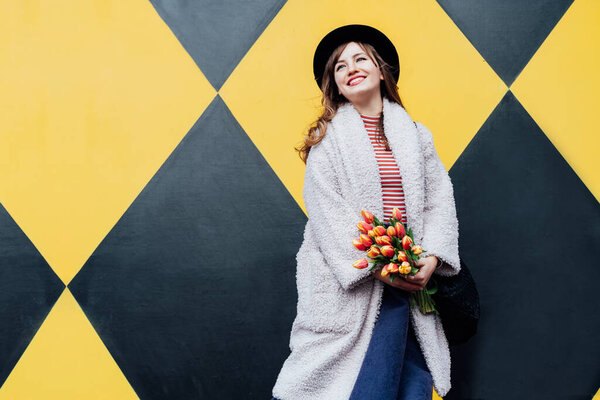 Happy. emotional young fashion woman holding bouquet of fresh tulip flowers on the geometry bright yellow and black wall background. Urban city street fashion. Spring mood, women day. Selective focus
