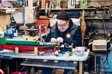 Emekli yaşlı bir adam garajındaki masada el işi gemi modeli yapıyor. Emekli adam elektrikli tornavidayla tekne modelini monte ediyor. Aktif emeklilik yaşam tarzı, hobi. Seçici odak