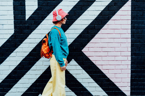 Side view stylish woman in bright clothes and bucket hat wearing wireless headphones and listening to music while walking near city urban painted wall. Fashionable hipster lifestyle. Selective focus