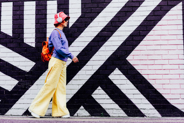 Side view stylish woman in bright clothes and bucket hat wearing wireless headphones and listening to music while walking near city urban painted wall. Fashionable hipster lifestyle. Selective focus