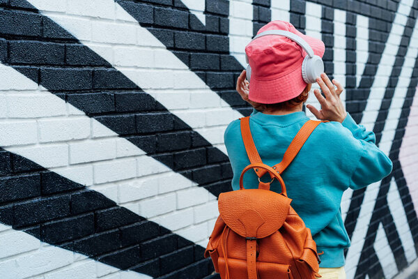 Back view stylish woman in bright clothes and bucket hat wearing wireless headphones and listening to music while walking near city urban painted wall. Fashionable hipster lifestyle. Selective focus