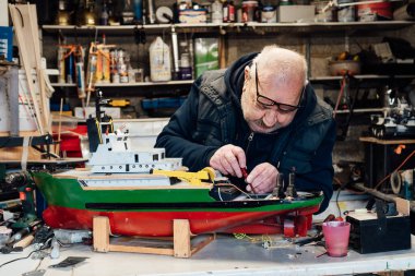 Emekli yaşlı bir adam garajındaki masada el işi gemi modeli yapıyor. Emekli adam tornavidayla tekne modelini monte ediyor. Aktif emeklilik yaşam tarzı, hobi. Seçici odak