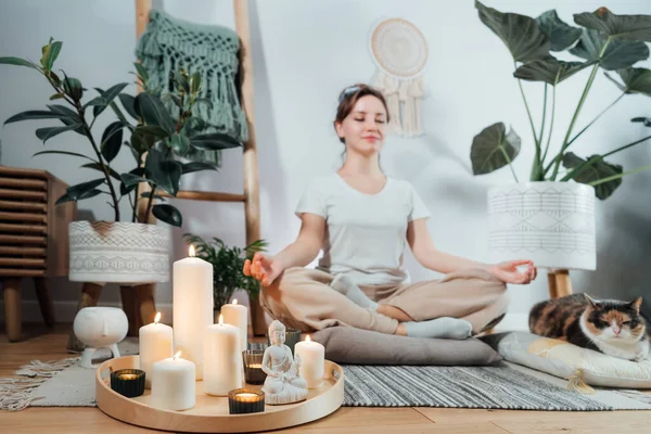 Focus on burning candles and white Buddha statuette on tray with background of woman practicing meditation at home with cat in modern Scandi interior. Zen Composition for yoga practice, relaxation