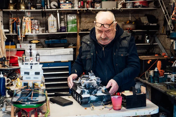 stock image Senior man, pensioner holding made handicraft scale model of a tank on the table ih his garage with different materials. Retired elderly man hobby. Active Retirement lifestyle. Selective focus.