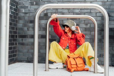 Hippi modasında, parlak elbiseli, güneş gözlüklü, kova şapkalı, sırt çantalı ve kulaklıklı genç bir kadın gri tuğla duvarın yanında oturuyordu. Şehir sokak modası. Seçici odak