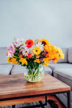 Kahve sehpasının üzerinde çeşitli çiçek buketi olan, gri kanepeli, modern, rahat, aydınlık oturma odasının bulanık arka planına sahip büyük bir vazo. Çiçek vermek. Anneler günü, doğum günü hediyesi.
