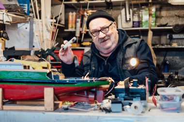 Emekli yaşlı bir adam garajındaki masada el işi gemi modeli yapıyor. Emekli adam elektrikli tornavidayla tekne modelini monte ediyor. Aktif emeklilik yaşam tarzı, hobi. Seçici odak.