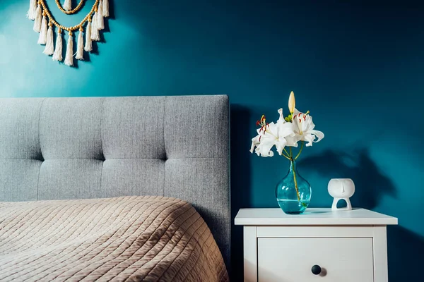 Cozy bedroom with turquoise blue walls and Boho home interior decor: macrame wall hanging decoration, white bedside table,vase with lilly flower, aroma lamp. Dark modern stylish room. Selective focus