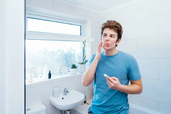 にきびの問題を持つ集中白人の十代の少年は自宅でケア顔の皮膚を取ります 彼は鏡を見て バスルームで顔にクリームを塗る 10代のスキンケア毎日の治療プロセス — ストック写真