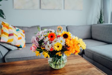 Kahve sehpasının üzerinde çeşitli çiçek buketi olan, gri kanepeli, modern, rahat, aydınlık oturma odasının bulanık arka planına sahip büyük bir vazo. Çiçek vermek. Anneler günü, doğum günü hediyesi.