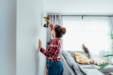 Ekose gömlekli genç esmer kadın elektrikli tornavidayla duvara vida sokuyor. Kız evdeki duvara resim asmak istiyor. Oda temizliği işi. Kendini tamir ediyor. DIY kavramı.