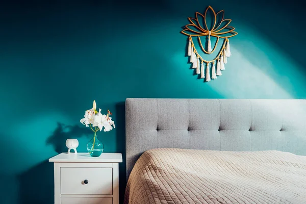 Cozy bedroom with turquoise blue walls and Boho home interior decor: macrame wall hanging decoration, white bedside table,vase with lilly flower, aroma lamp. Dark modern stylish room. Selective focus