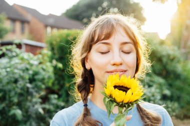 Close up portrait of tender young woman with closed eyes in blue t shirt holding fresh yellow sunflower and enjoying the moment in sunset light. Summertime. Selective focus