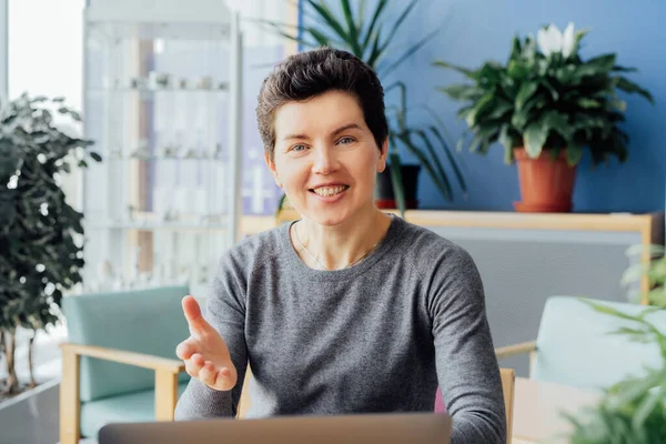 Smiling neutral gender middle aged woman with no make-up in casual clothing emotionally talking and looking at camera at her workstation in an open space office. Online video call portrait
